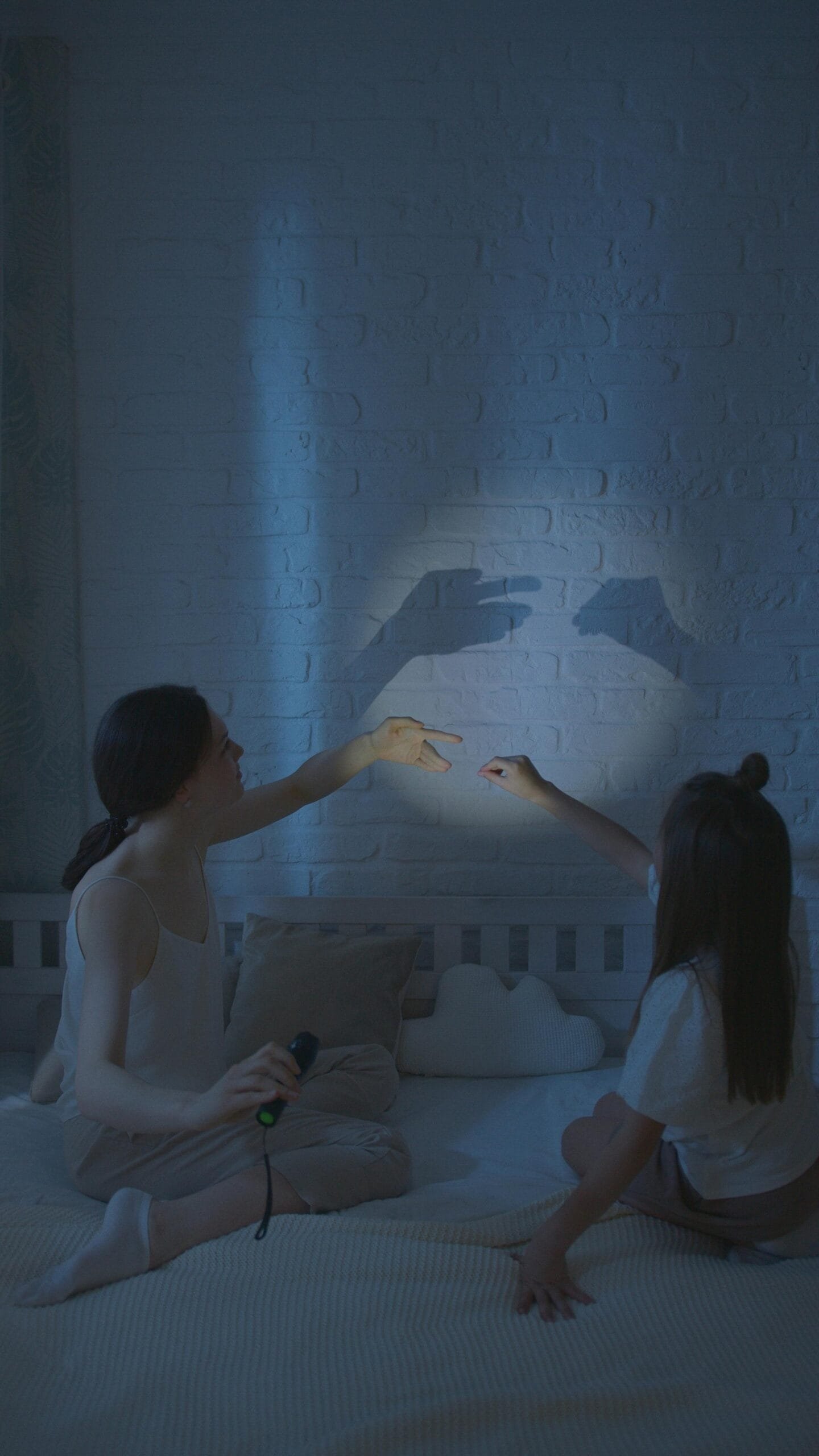 A mother and daughter create shadow puppets on a bedroom wall, enjoying a nighttime play session.